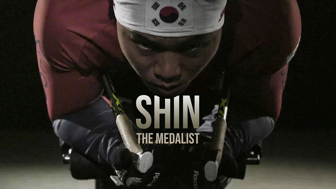 A male athlete in a white headband with South Korean flag and Taegeuk symbol, wearing a red shirt, leaning forward on rowing oars in a dark setting. Text overlay says "SHIN THE MEDALIST."