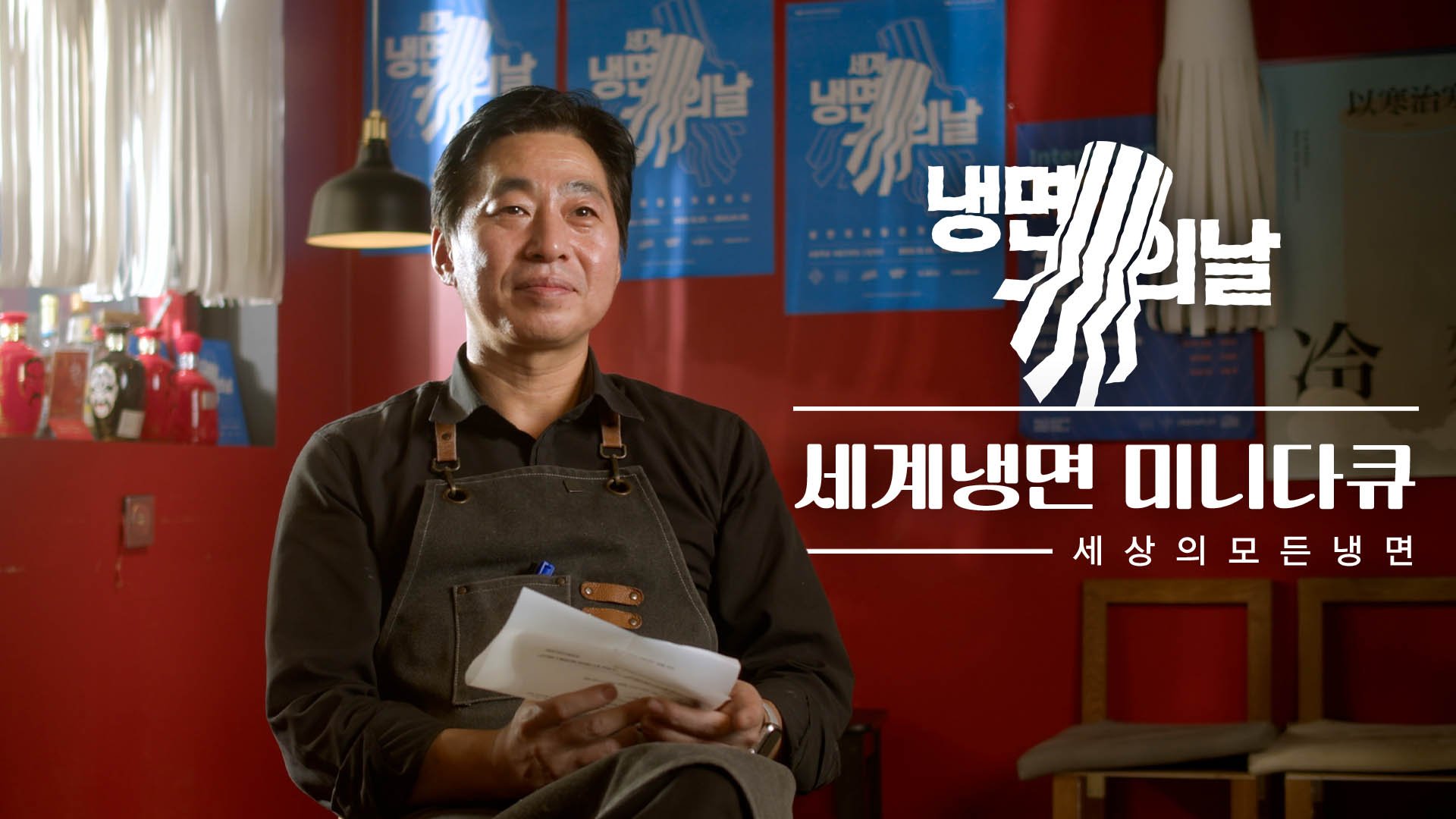 Man with black hair and apron, sitting with legs crossed, holding papers, in a red room with bottle displays, and blue posters behind him. Korean text overlay.