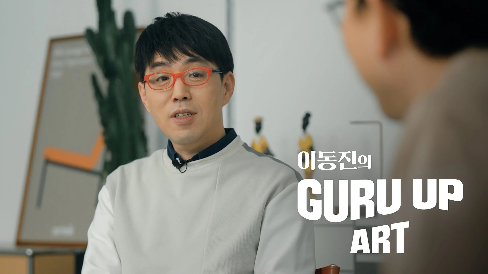 A man with black hair and red glasses speaks during an interview or conversation, with Korean and English text overlaying on the right side that reads 'GIRL UP' and 'ART'. In the background, there's a tall cactus plant and a framed artwork or poster on the wall.