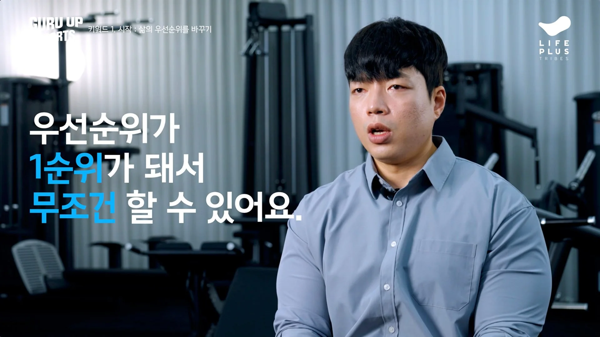 A man in a gray shirt sitting in an office or conference room with equipment and window blinds in the background, with Korean text overlaying the image.