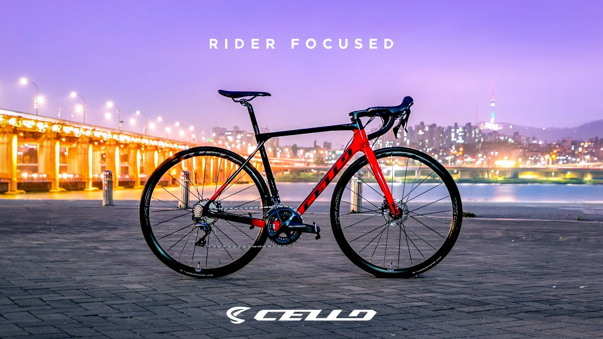 A Trek road bicycle black and red, parked on a brick paved area near a city river at dusk with city lights and a bridge in the background, with the word 'CERVELO' on the ground in the foreground.