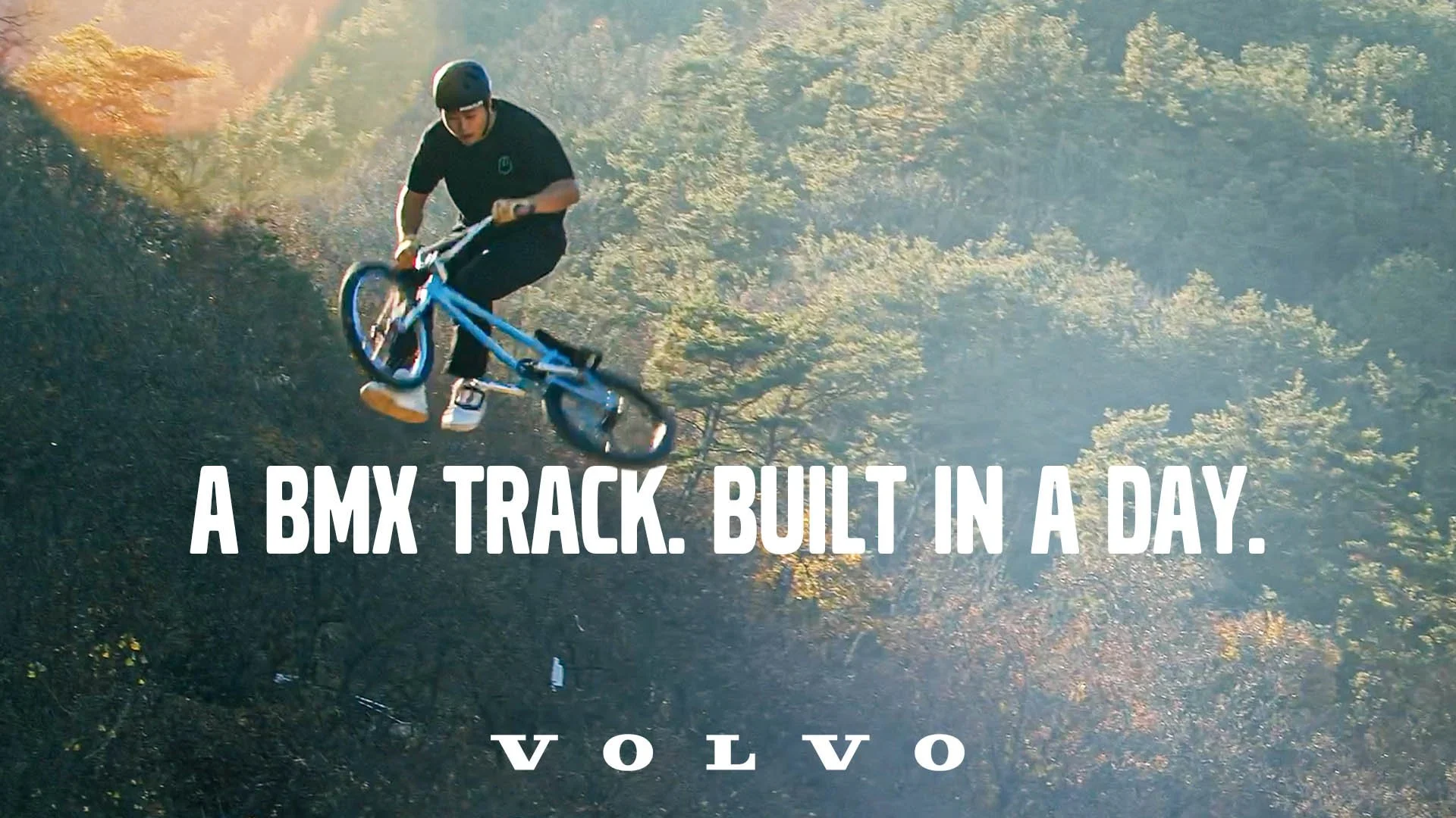 A person wearing a helmet riding a BMX bike on a dirt track in a wooded area with trees and sunlight.