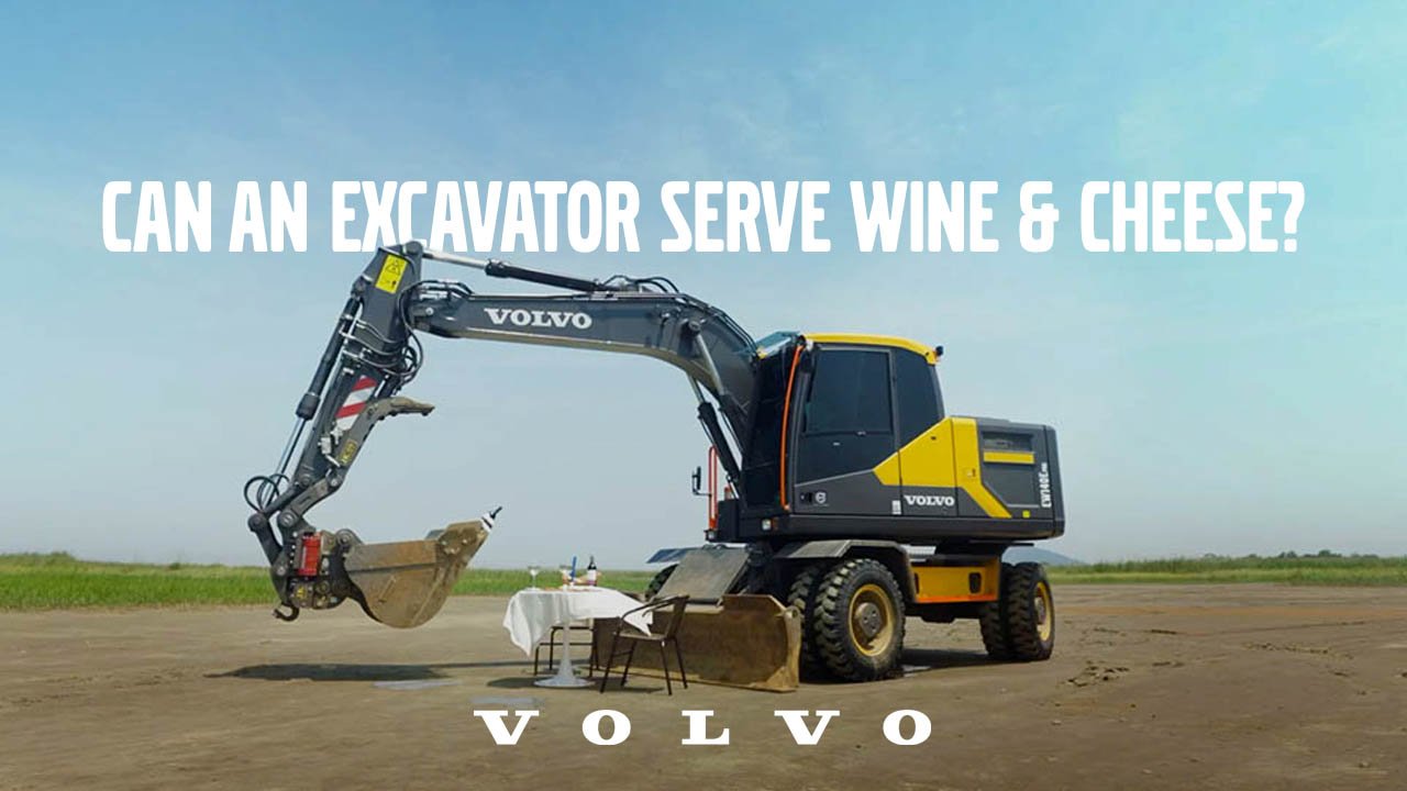 A large Volvo excavator with an open bucket, positioned in an open field under a blue sky, appears to be serving wine and cheese from a table placed in front of it.