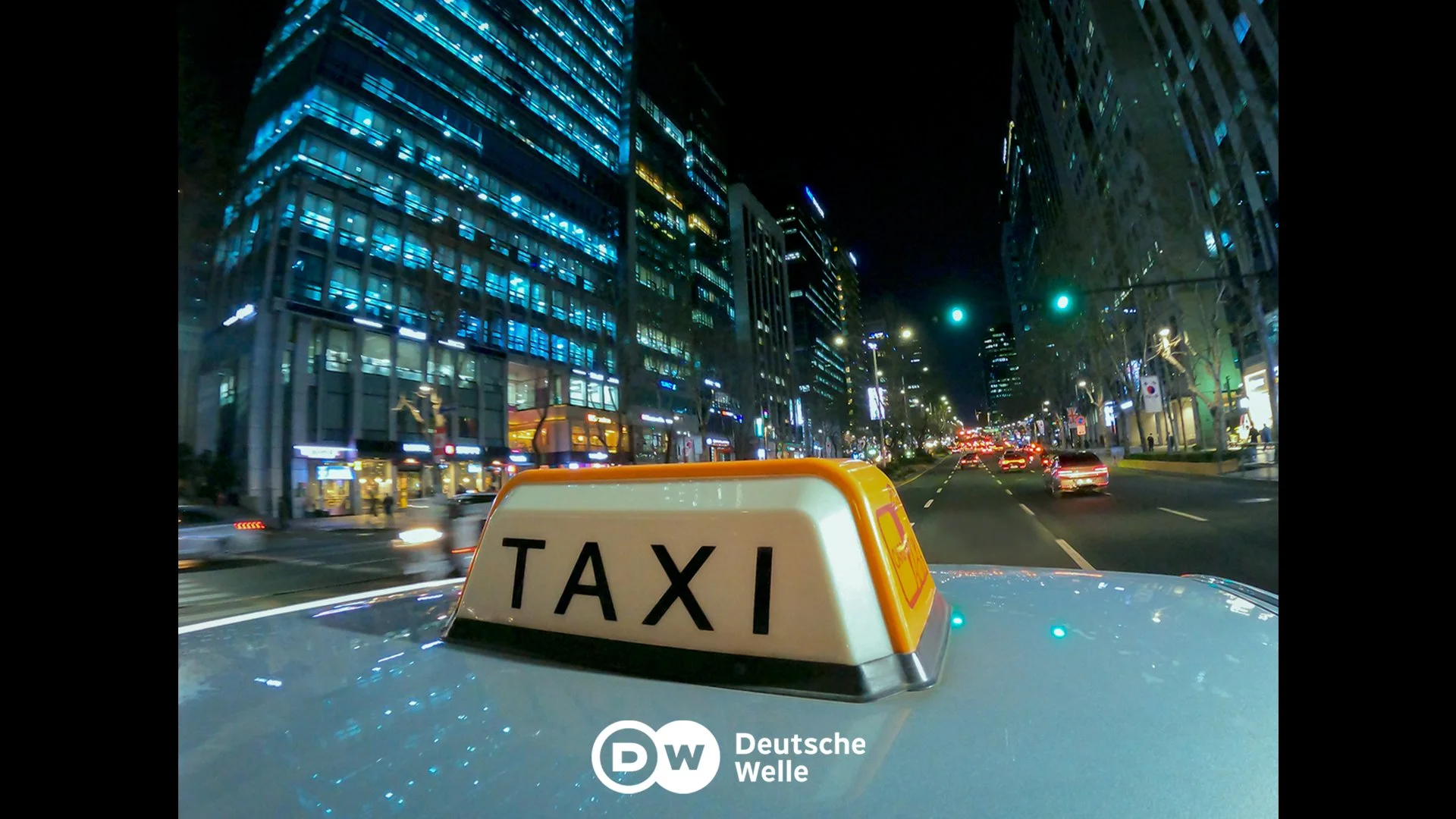 Nighttime city street viewed from the front of a taxi with a taxi sign on the roof. Tall buildings with illuminated windows line both sides of the street. Traffic is moving along the road, and there are traffic lights overhead. The scene is brightly lit with city lights and reflections on the buildings.