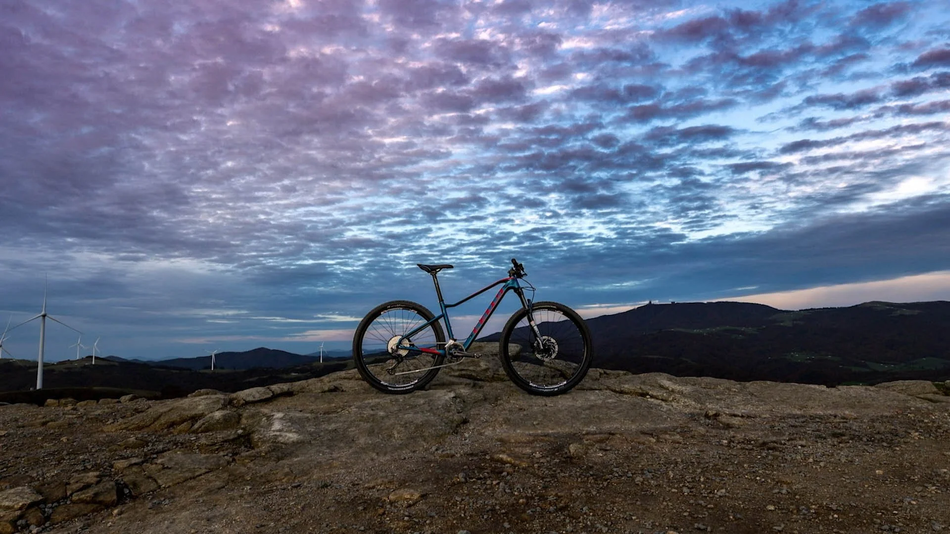 A mountain bike stands on rocky terrain against a backdrop of rolling hills and a cloudy sky at dusk with wind turbines visible in the distance.