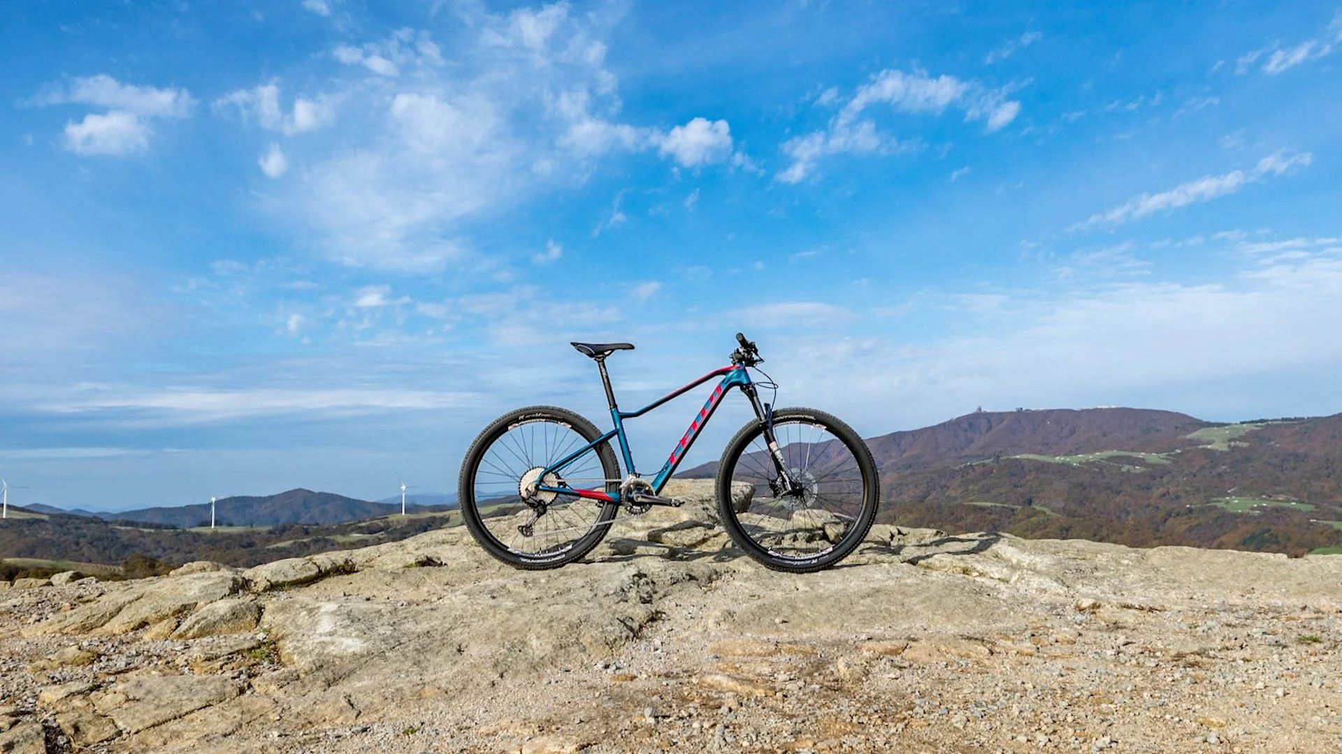 A mountain bike on rocky terrain with wind turbines and mountains in the background under a blue sky with clouds.