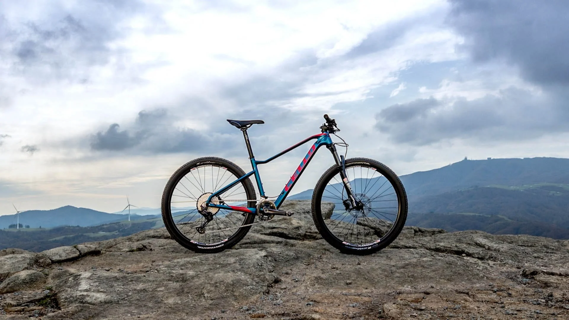A mountain bike resting on a rocky outcrop with a scenic landscape of rolling hills and wind turbines visible in the distance under a cloudy sky.