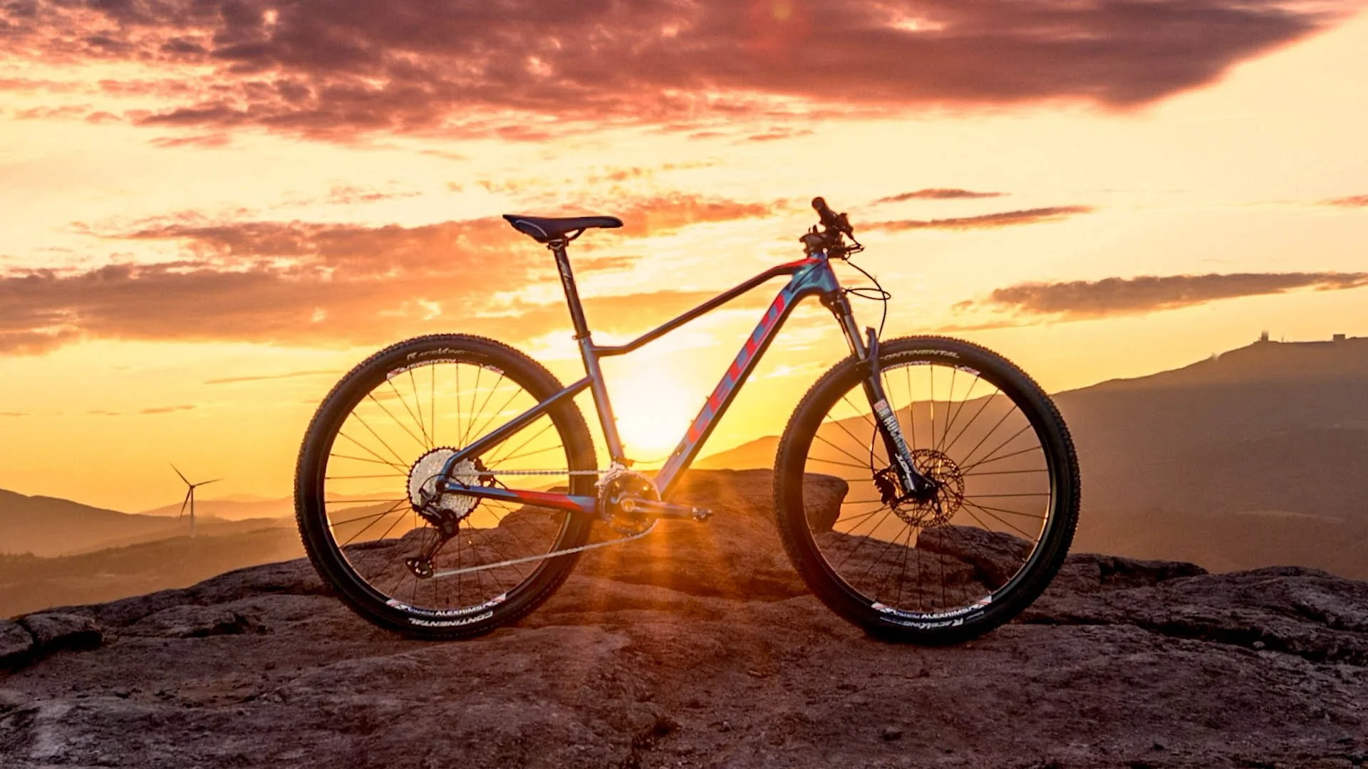 A mountain bike standing on rocky terrain during a sunset with a colorful sky, mountain silhouette, and a wind turbine in the background.