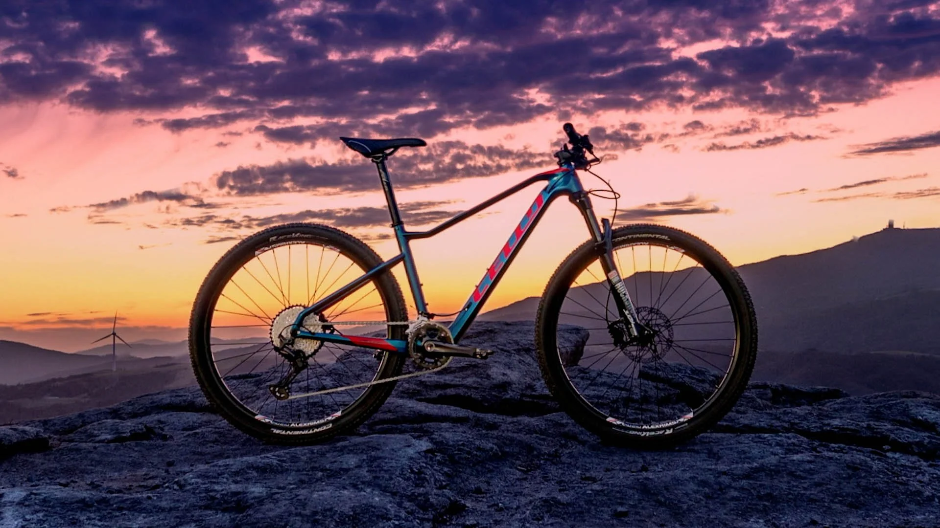 A mountain bike on a rocky surface at sunset with pink and purple clouds, distant mountains, and a wind turbine in the background.