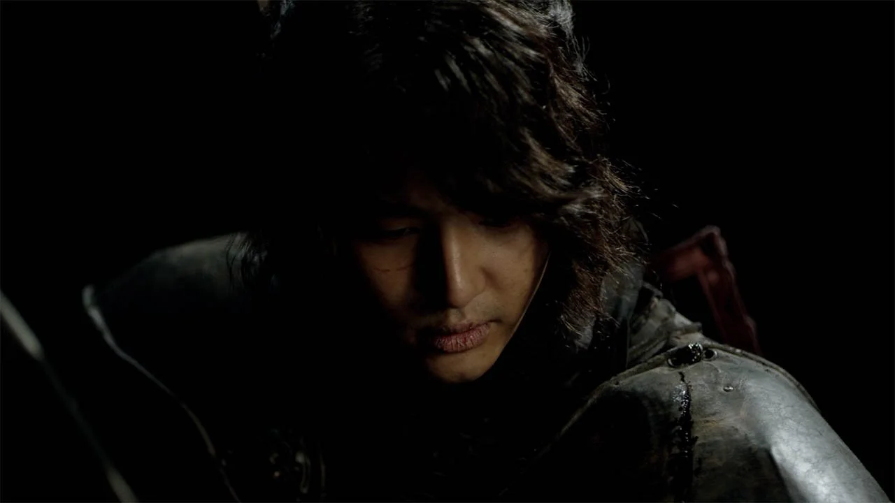 A person with long, wavy dark hair and light skin with a serious expression, wearing a dark outfit with metallic details, in a low-light setting.