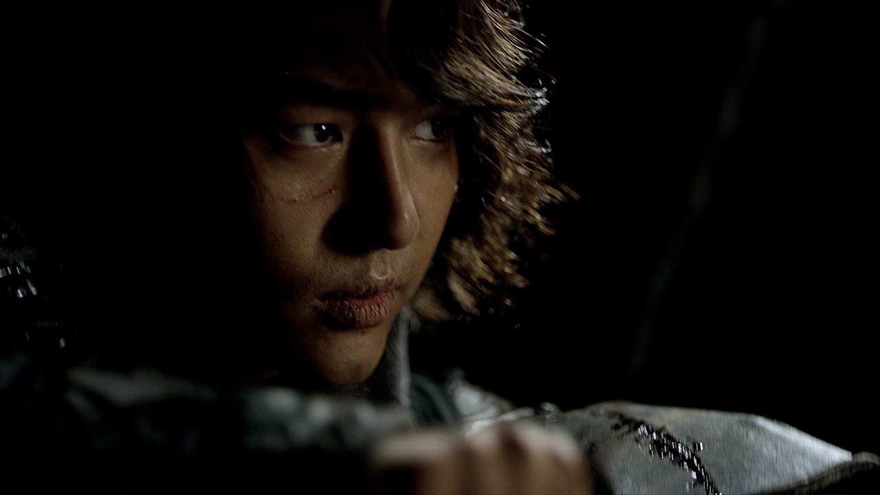 A close-up of a person with light brown hair and a scar on their face, looking intently to the side in a dark setting.
