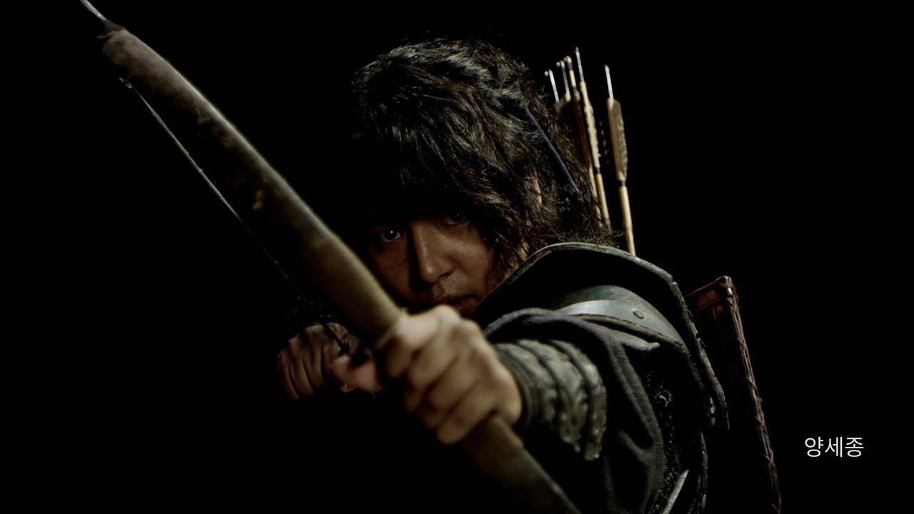 A person with dark, tousled hair and armor, holding a bow and aiming with intensity, with a quiver of arrows on their back against a dark background.