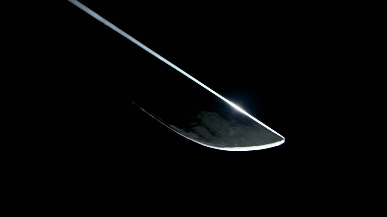Close-up of a sharp, curved knife blade with light reflecting off the edge against a black background.
