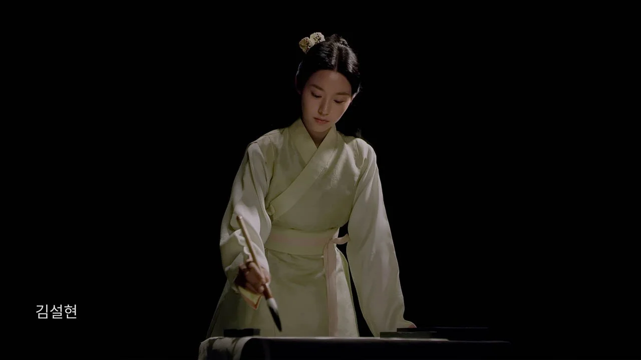 A woman dressed in traditional Korean hanbok, holding a calligraphy brush over a table, with a black background. Text in Korean is at the lower left corner.