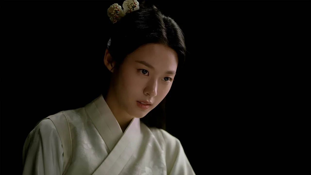 Young woman with traditional Korean attire and ornamented hair, looking contemplative, against a black background.