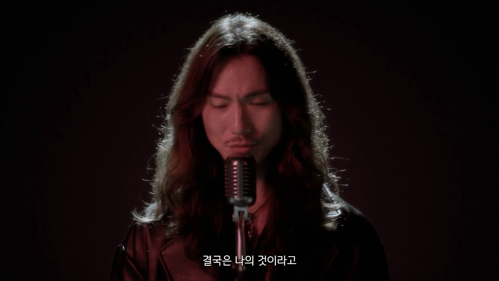 A person with long wavy hair singing into a vintage microphone against a dark background, with Korean subtitles at the bottom.