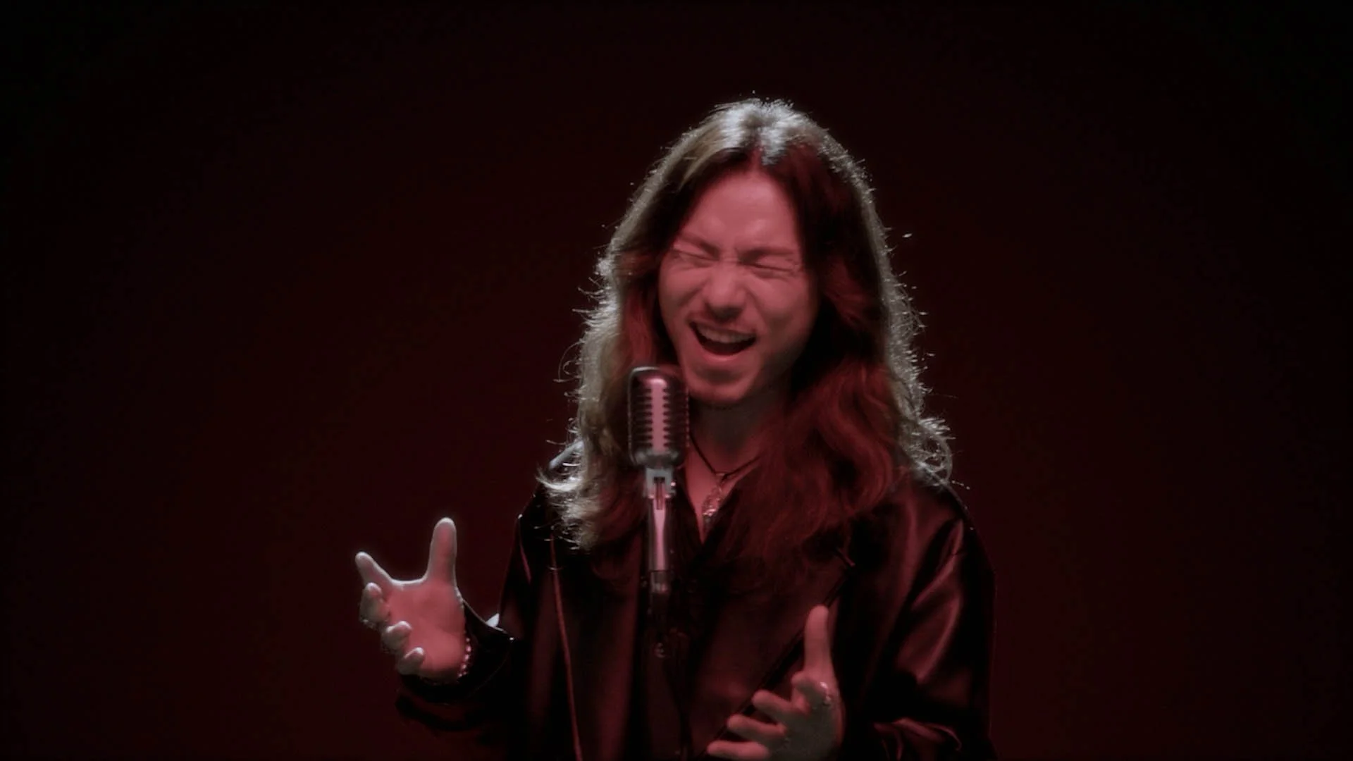 A man with long hair, singing passionately in front of a vintage microphone, wearing a shiny black jacket, with tears and emotion visible on his face.
