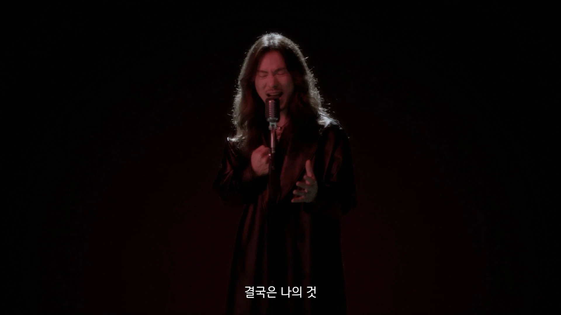 Person singing passionately into a vintage microphone on a dark stage with Korean subtitles at the bottom.