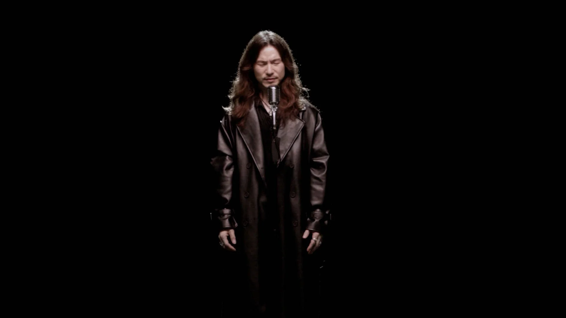 A man with long hair and a leather trench coat singing into a vintage microphone against a black background.
