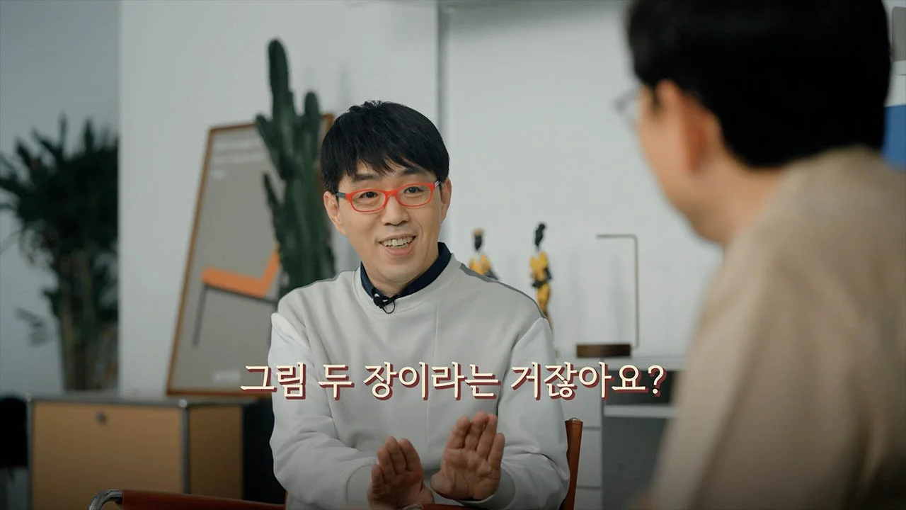 A man with black hair and red glasses smiling and talking to another person in an indoor setting with large green plants in the background.