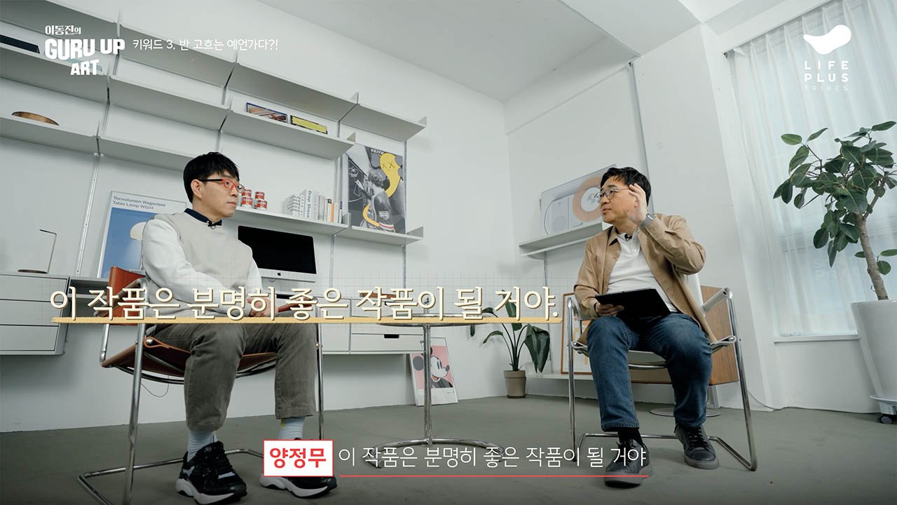 Two men sit in a bright, minimalist office engaged in a conversation. The man on the left wears glasses, a white shirt, and gray pants, while the man on the right wears glasses, a tan jacket, and jeans. There are books, plants, and decorative items on shelves behind them, with a window with sheer curtains on the right.