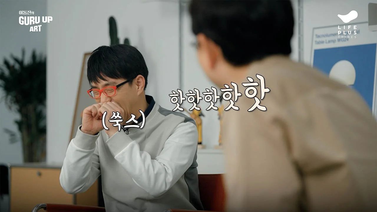 Two people in an office, one with black hair and red glasses, laughing with hand covering mouth, the other with dark hair, wearing a beige jacket, facing away. Korean text overlay, plants and a poster visible in background.