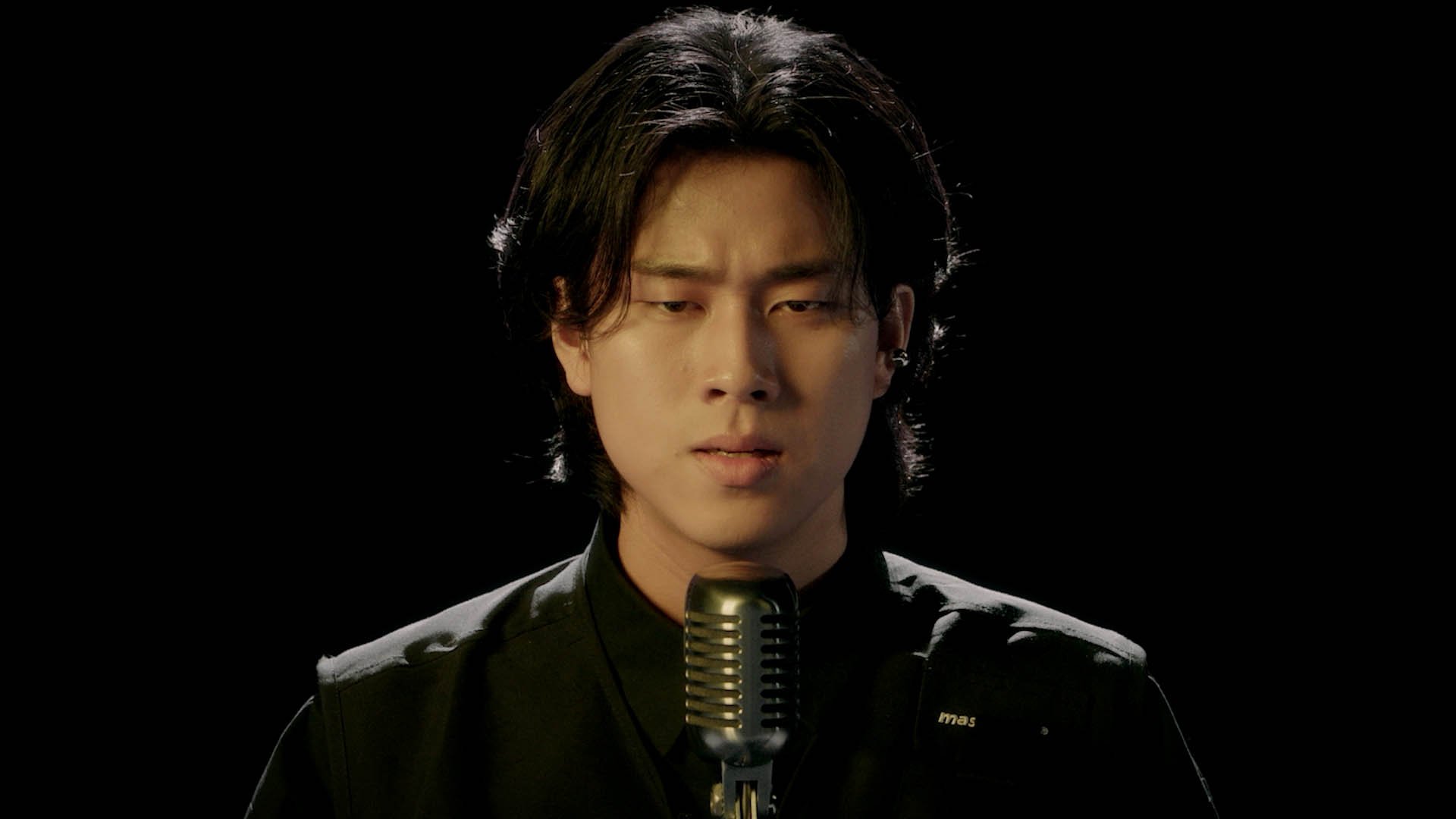 A young man in a black suit performs a vocal act with a vintage microphone against a dark background.