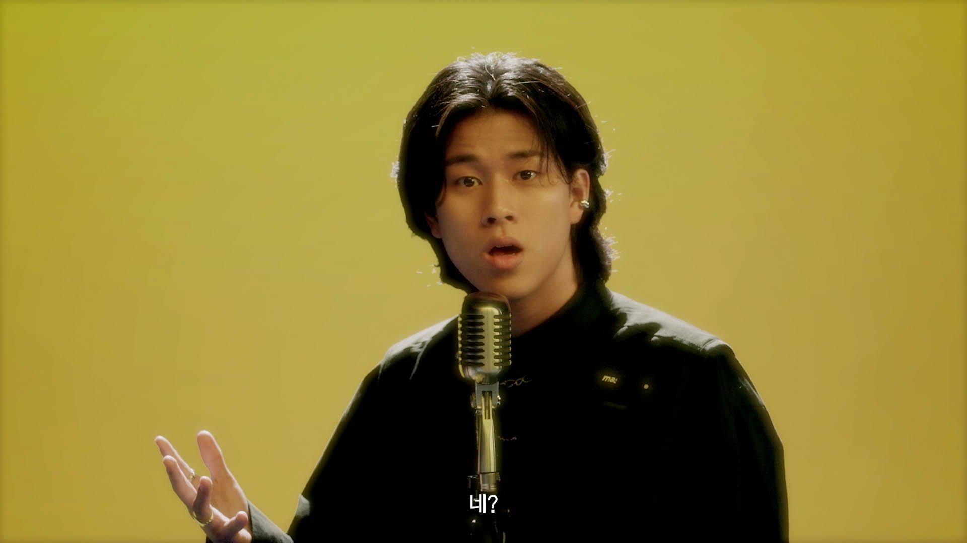 Young man singing into a vintage microphone against a yellow background, with Korean subtitles at the bottom.