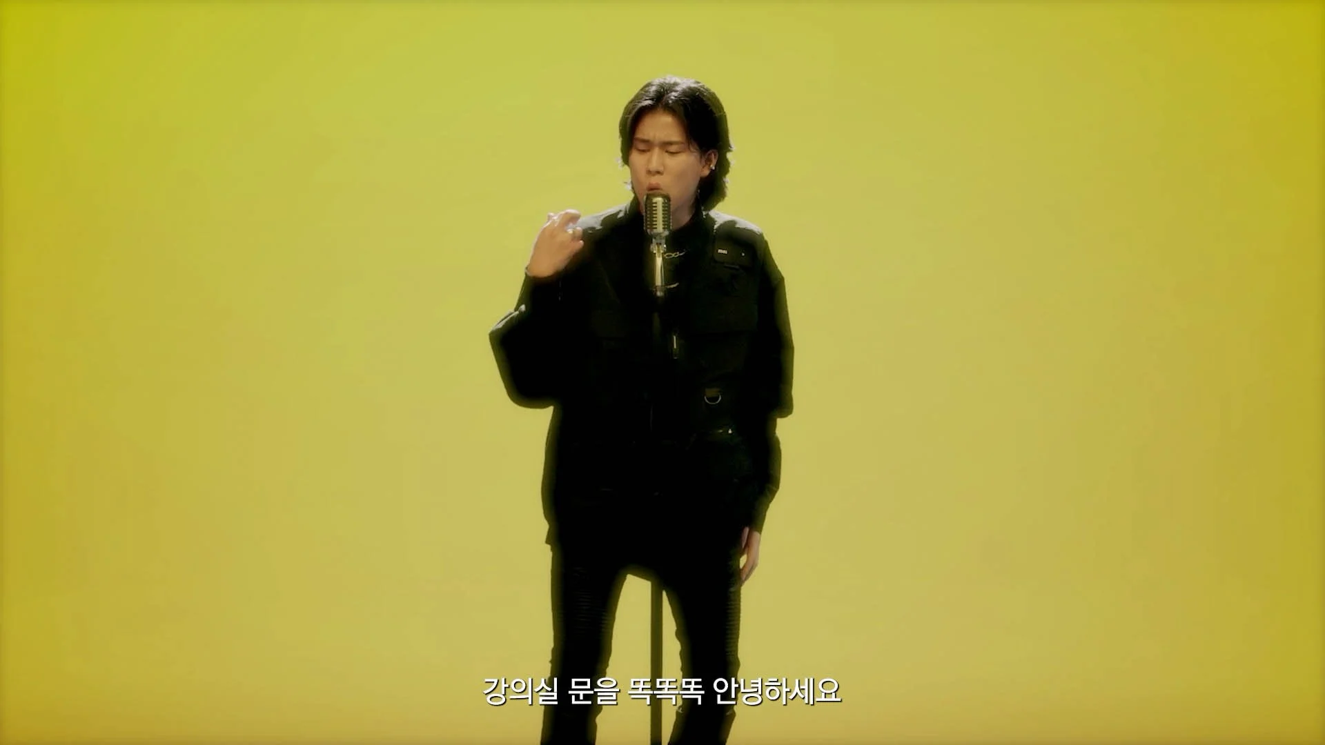A person with dark hair wearing a black outfit, standing in front of a yellow background, singing into a vintage microphone.