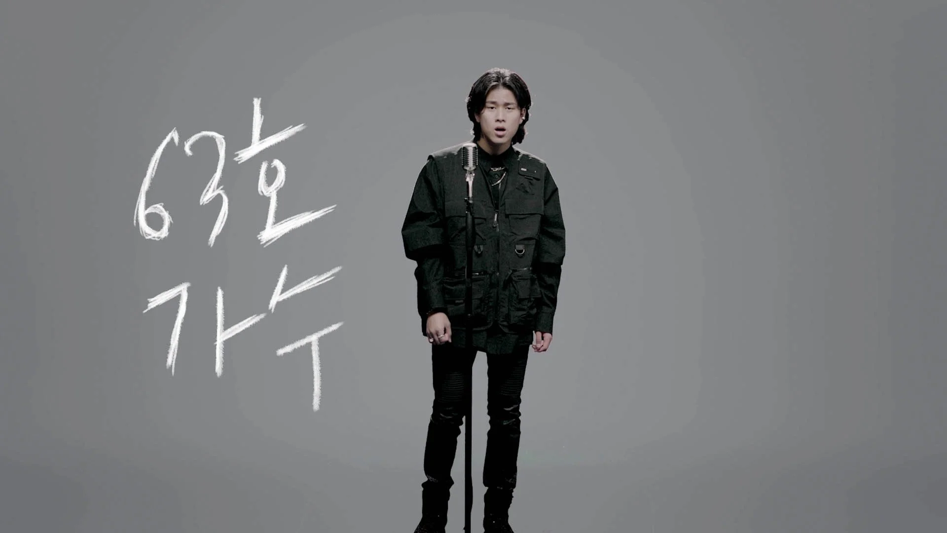A young man with black hair singing into a microphone against a gray background, with Korean characters written in white beside him.