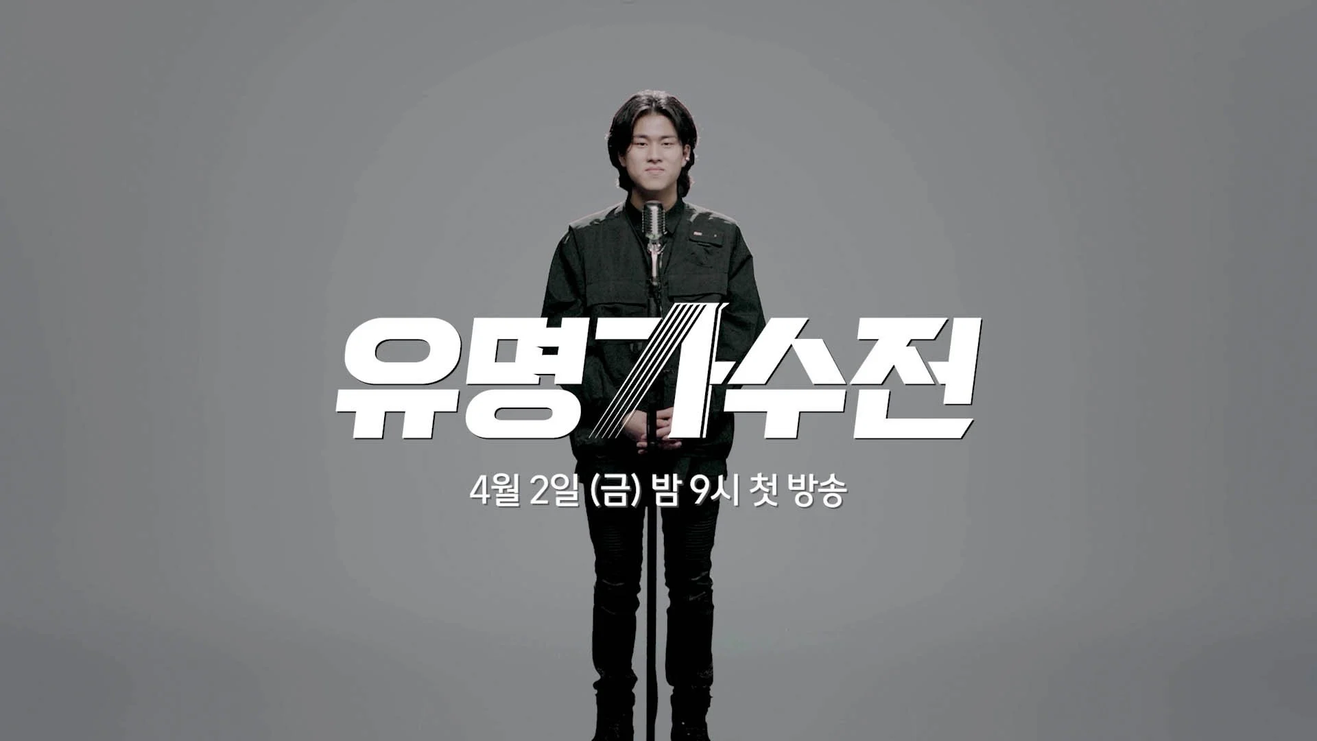 A person with short black hair and a black outfit standing in front of a microphone with a plain gray background. There is Korean text on the image.