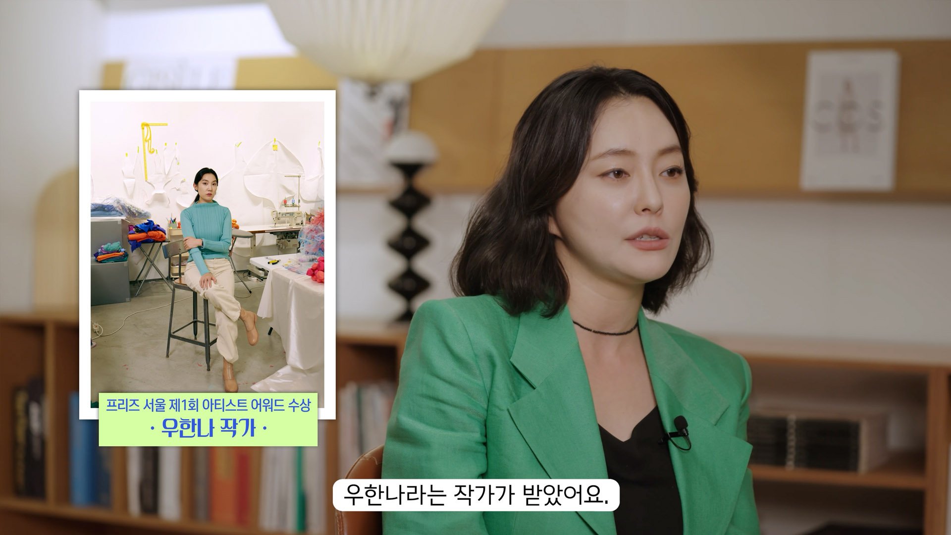 A woman with black wavy hair wearing a green blazer and black shirt, speaking during an interview in a room with bookshelves and a lamp. An inset image shows her sitting in a room with art supplies, fabrics, and a sewing machine, with Korean text at the bottom.