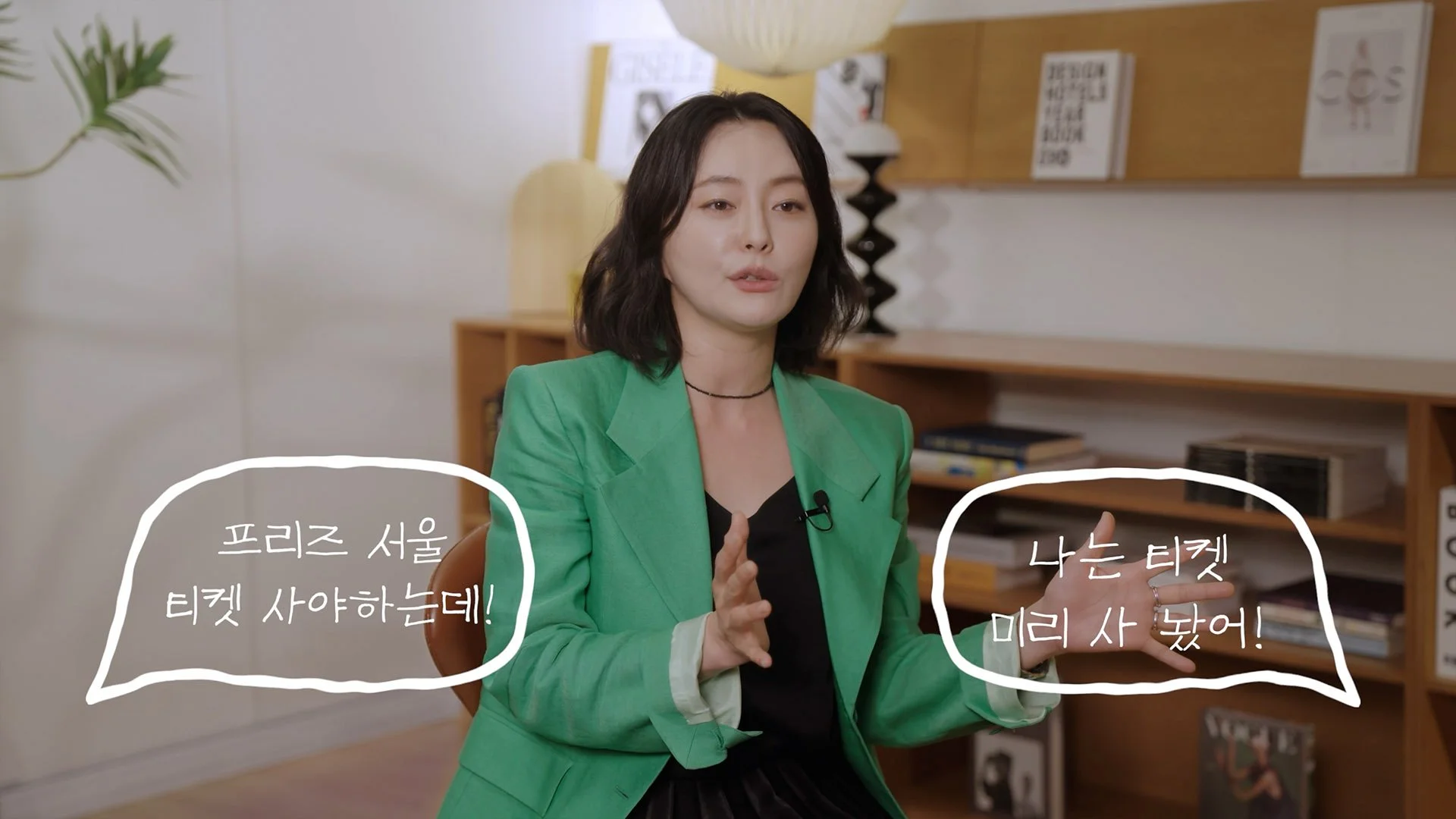 A woman with shoulder-length dark hair, wearing a bright green blazer, sits in a room with bookshelves and wooden decor. She is gesturing with her hands while speaking. There are handwritten speech bubbles on the image with Korean text.