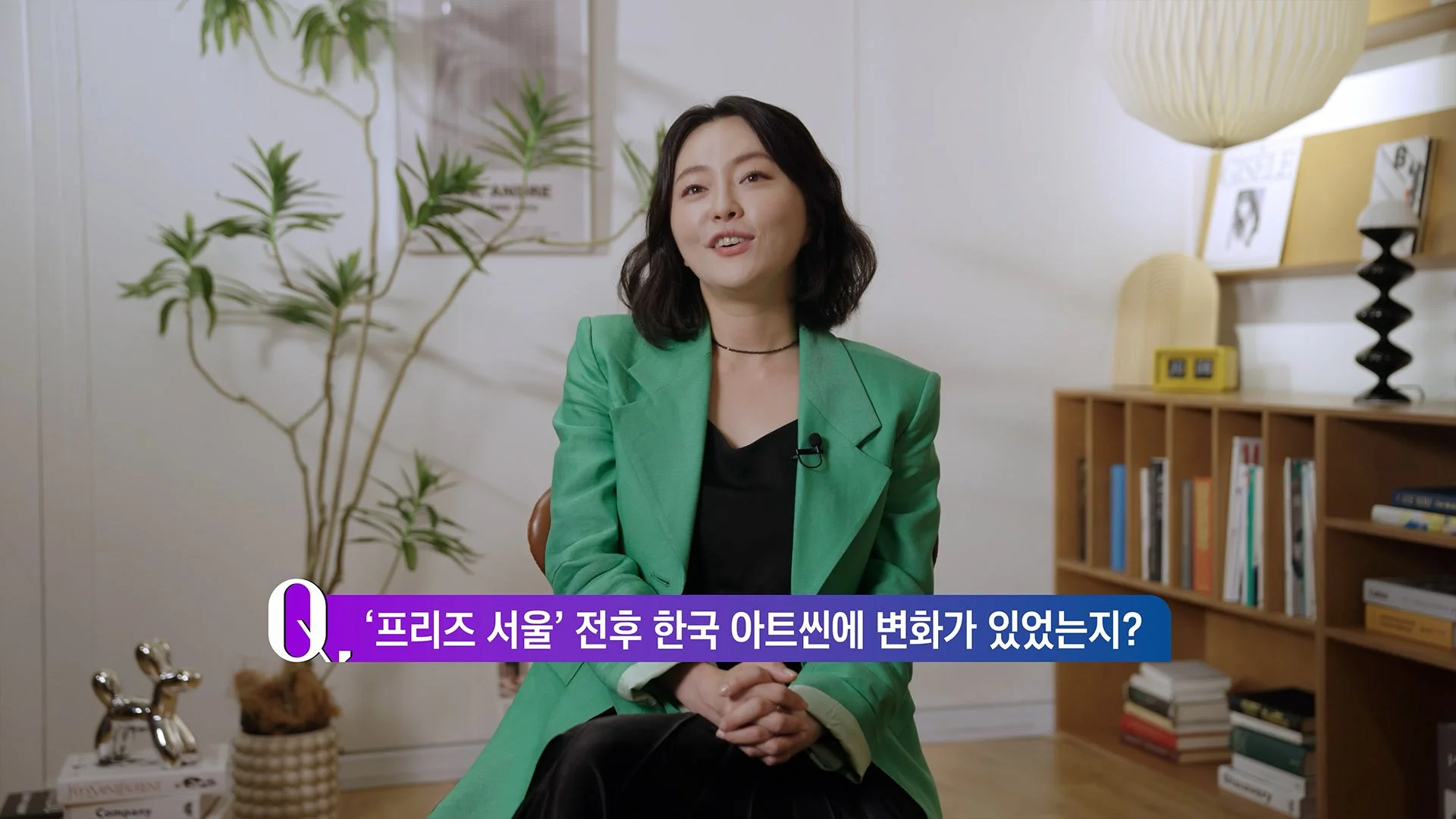 A woman with shoulder-length black hair, wearing a green blazer over a black top, sitting in a room with modern decor. There is a potted plant in the foreground and a wooden bookshelf with books behind her. A lamp and some framed pictures are also visible on the wall. The woman is engaged in conversation with a slight smile, and Korean text appears on the screen.