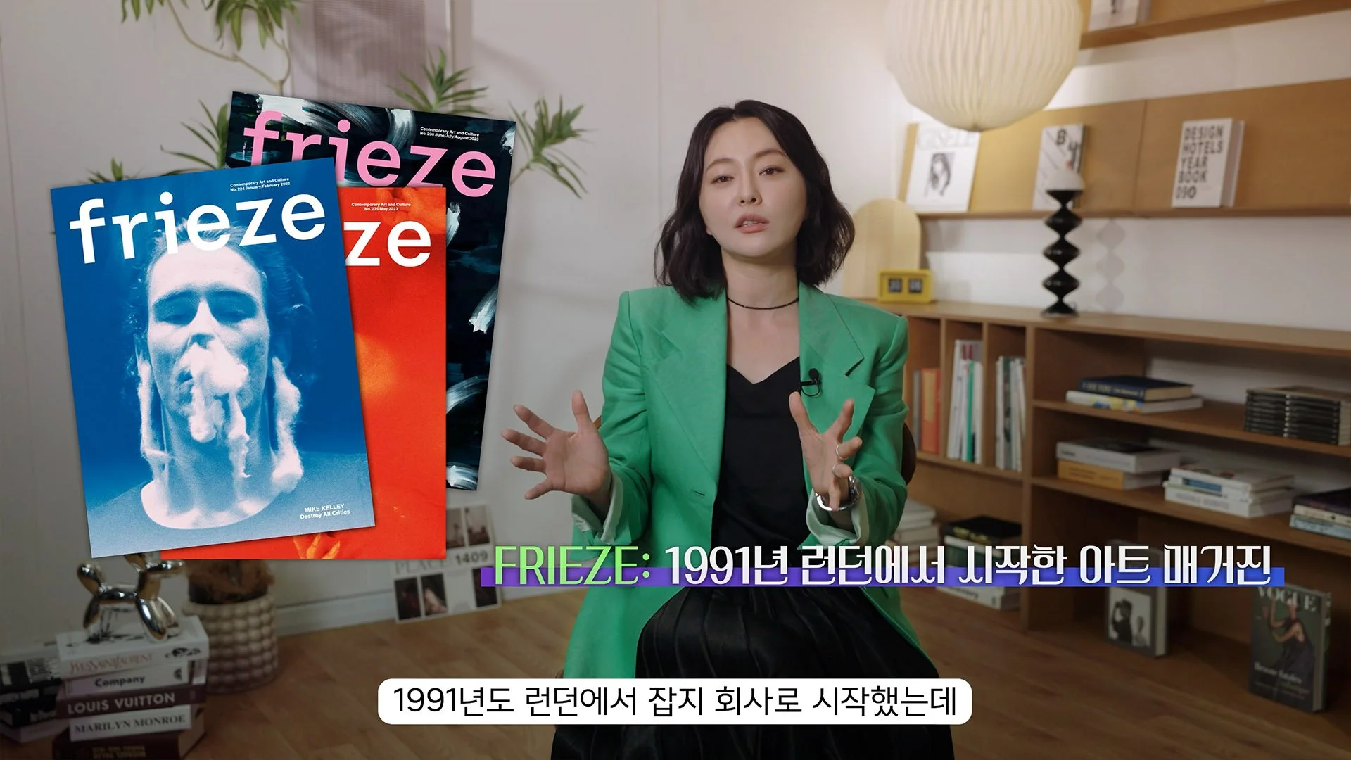 A woman with shoulder-length black hair wearing a green blazer and black dress, seated indoors in front of a wooden bookshelf and wall decorations. To her left, there are three magazines or posters titled "freeze" featuring a blue and white image of a person's face with steam or smoke coming from their nose. The woman appears to be speaking, with subtitles in Korean at the bottom of the image.