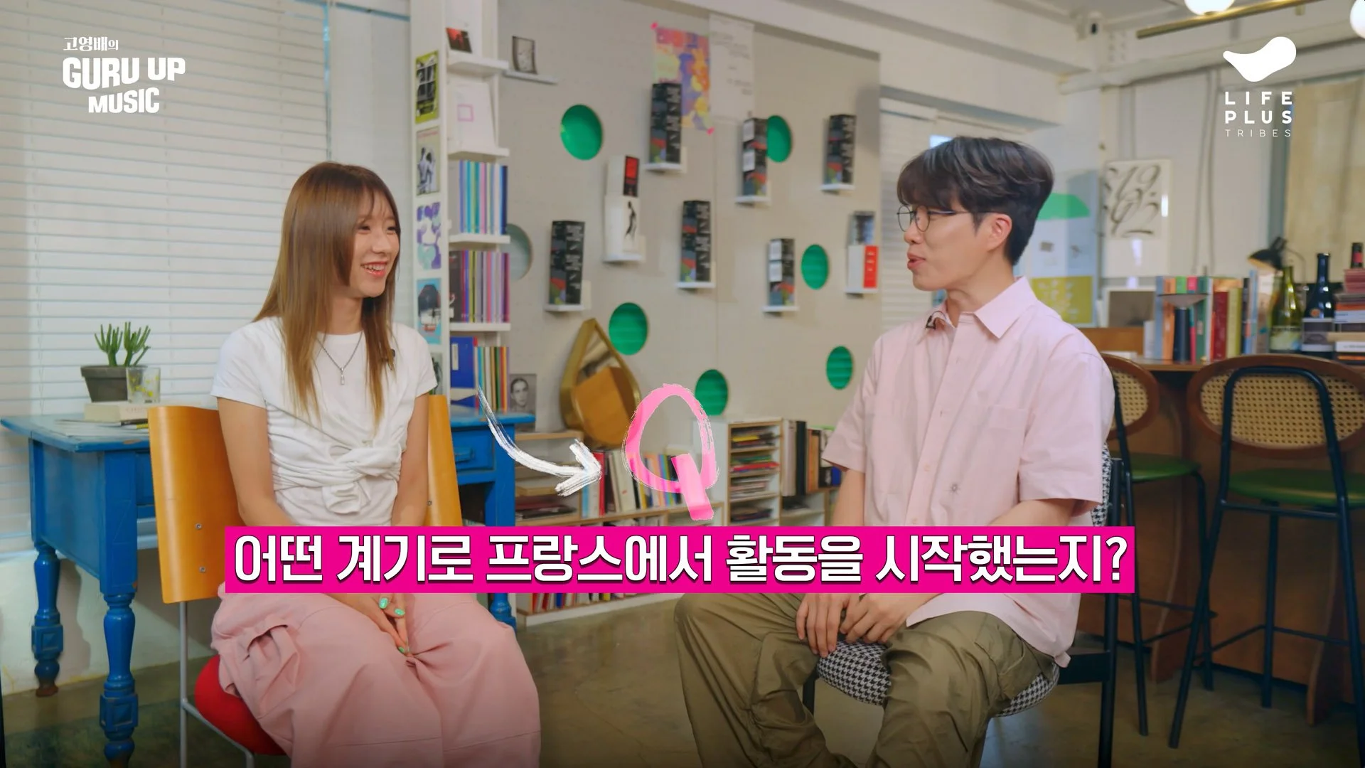 A woman and a man are sitting and talking in a bright, colorful room with bookshelves and artwork. The woman has long hair, is wearing a white t-shirt and pink pants, and is smiling. The man has glasses, dark hair, and is wearing a pink button-up shirt and khaki pants; he is speaking. There is Korean text at the bottom of the image with an arrow pointing to the woman.