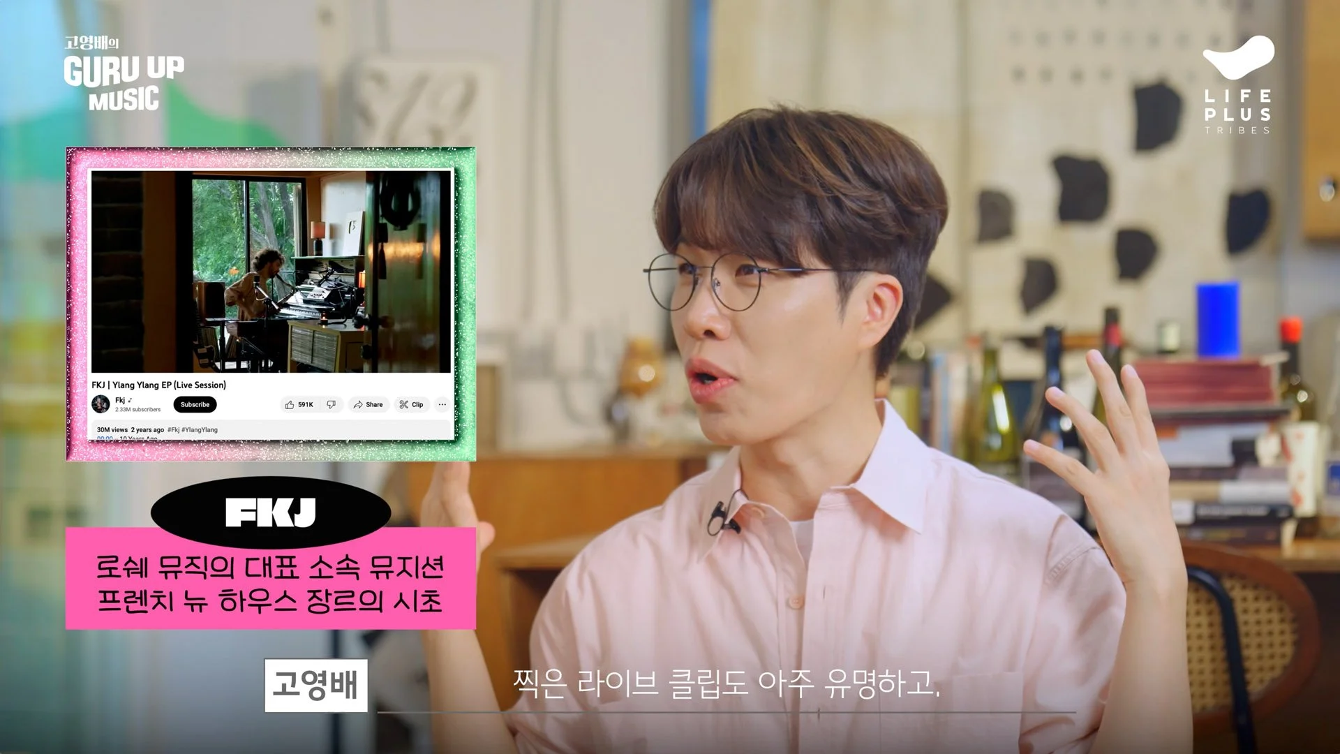 A man with glasses and brown hair, wearing a light pink shirt, is speaking while gesturing with his hand. In the foreground, a digital overlay shows a YouTube video thumbnail of a recording studio with a musician playing piano. The overlay includes text in Korean and a logo for 'FJK'. In the background, a classroom or studio setting is visible with shelves and various objects.