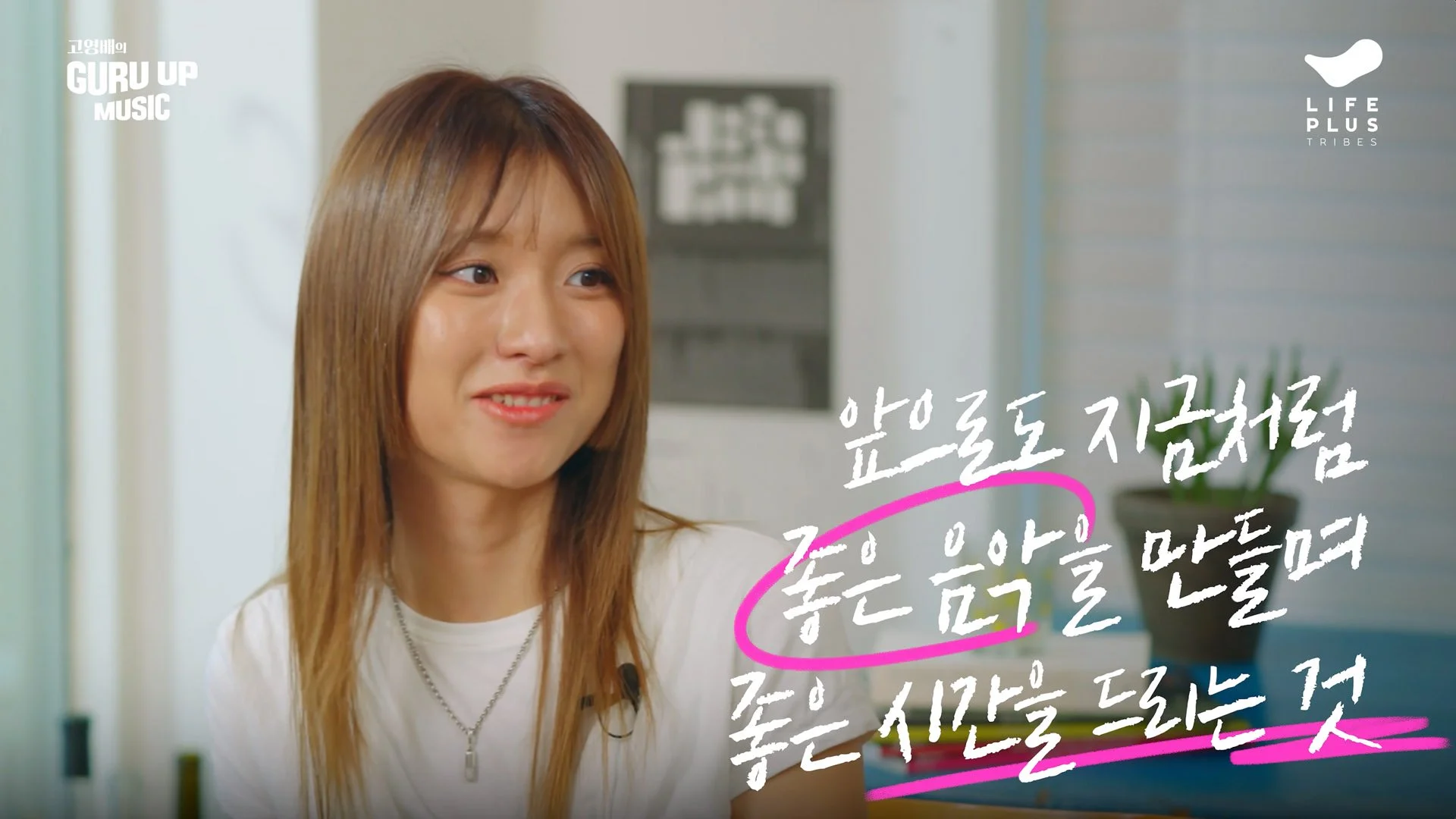 A woman with brown hair smiling, sitting indoors with Korean text overlaid on the image and a white wall in the background.