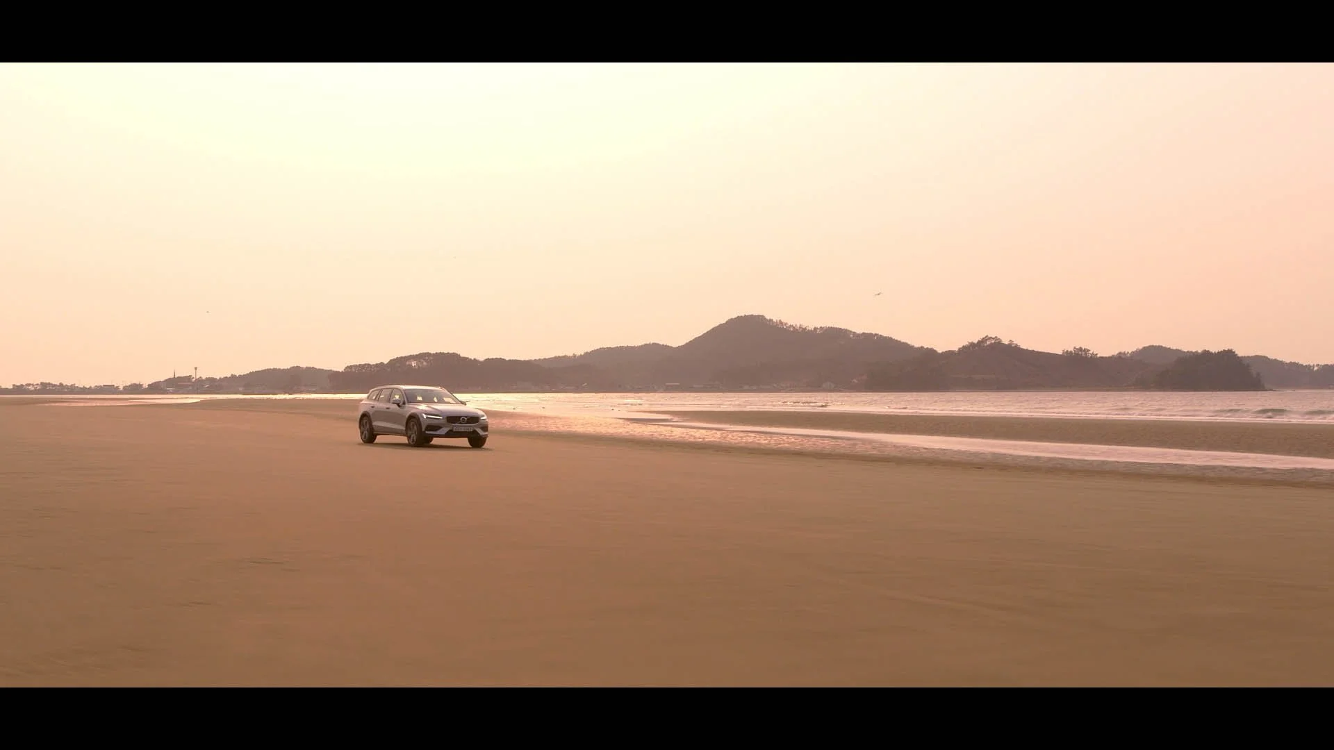 A silver SUV driving on a sandy beach with hills and water in the background during sunset or sunrise.