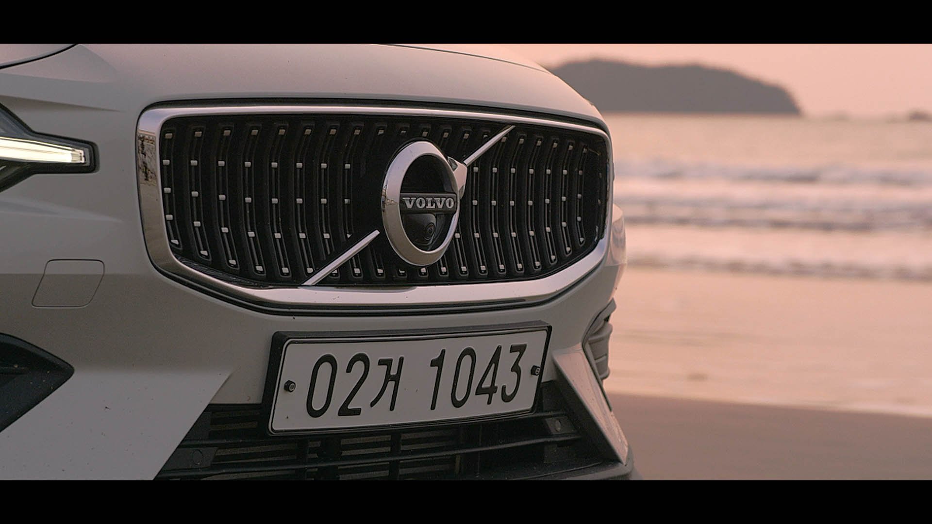 Front view of a white Volvo car parked near the beach during sunset, displaying its grille, headlights, and license plate.