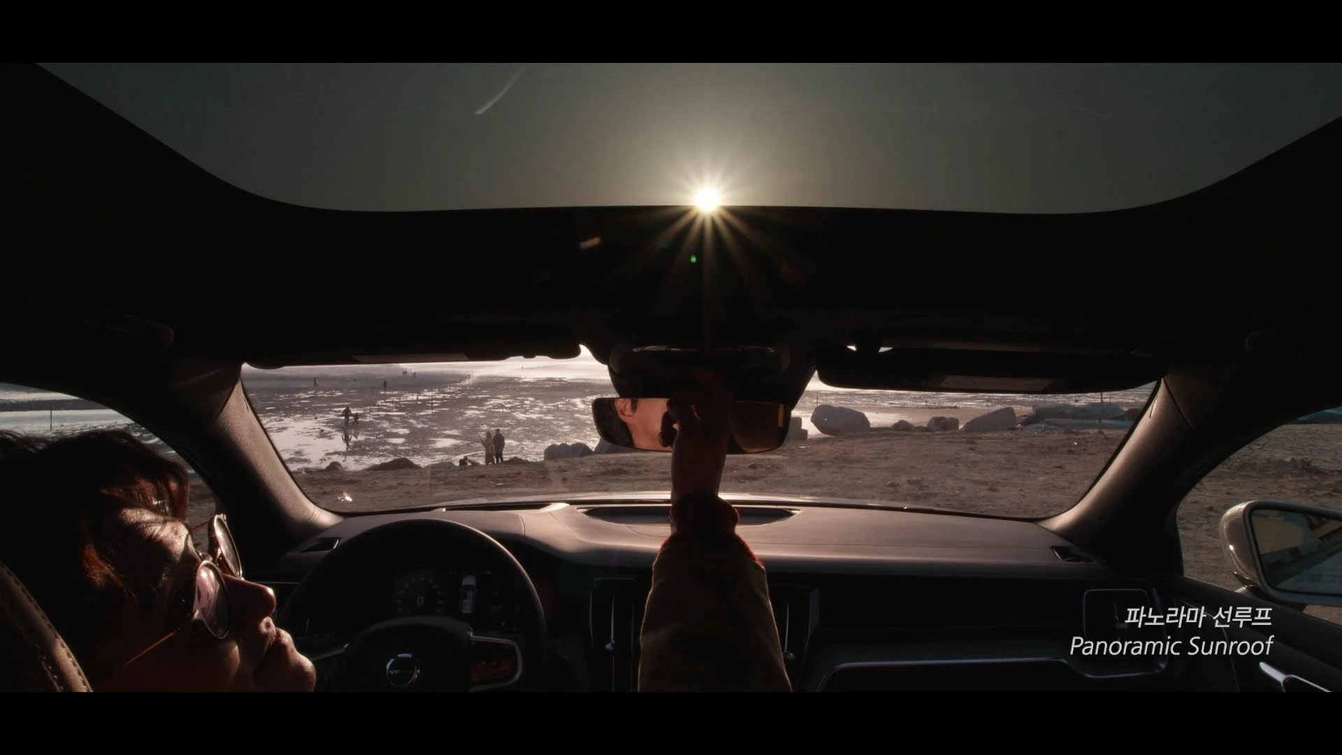 Inside a car at sunset with two passengers, one adjusting the rearview mirror overlooking a snowy landscape with a few people walking in the distance.