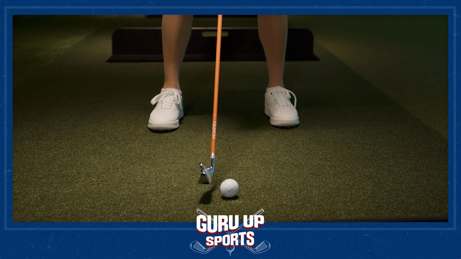 Person preparing to golf indoors, wearing white shoes, holding an orange golf club, with a golf ball on the dark green carpeted floor.