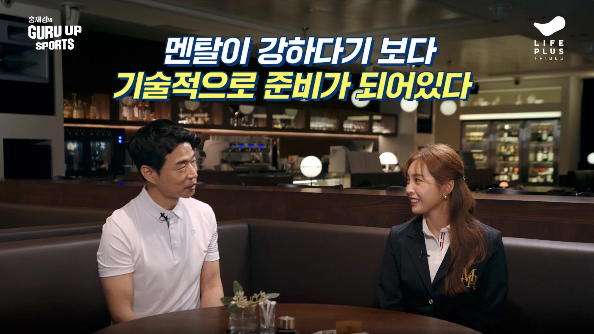 A man and a woman are seated at a table in a restaurant or cafe, having a conversation. The man is wearing a white shirt, and the woman is dressed in a dark blazer with a white shirt underneath. There is a plant in a small vase on the table, and the background features a modern bar or kitchen area with shelves and bottles.