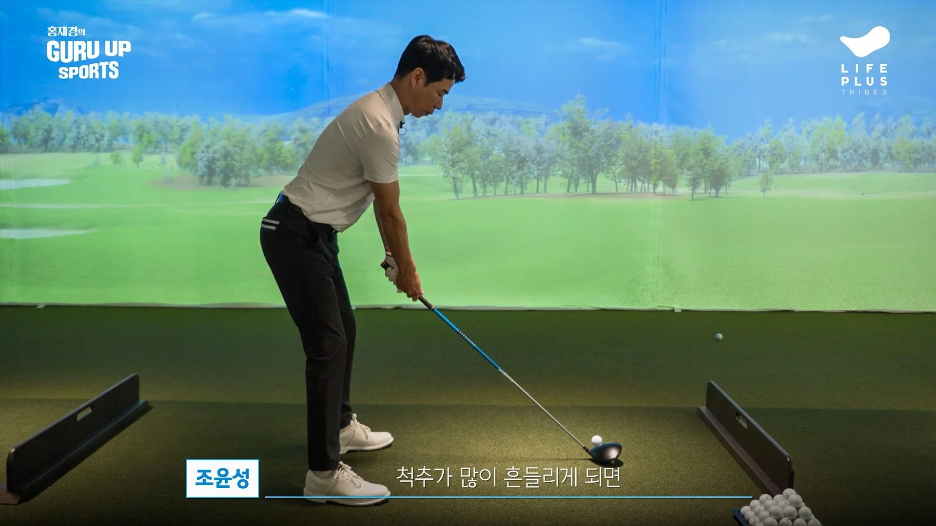 A man practicing golf indoors, preparing to hit the golf ball. The background displays a large screen with an outdoor golf course scene. There is Korean text and logos on the screen.
