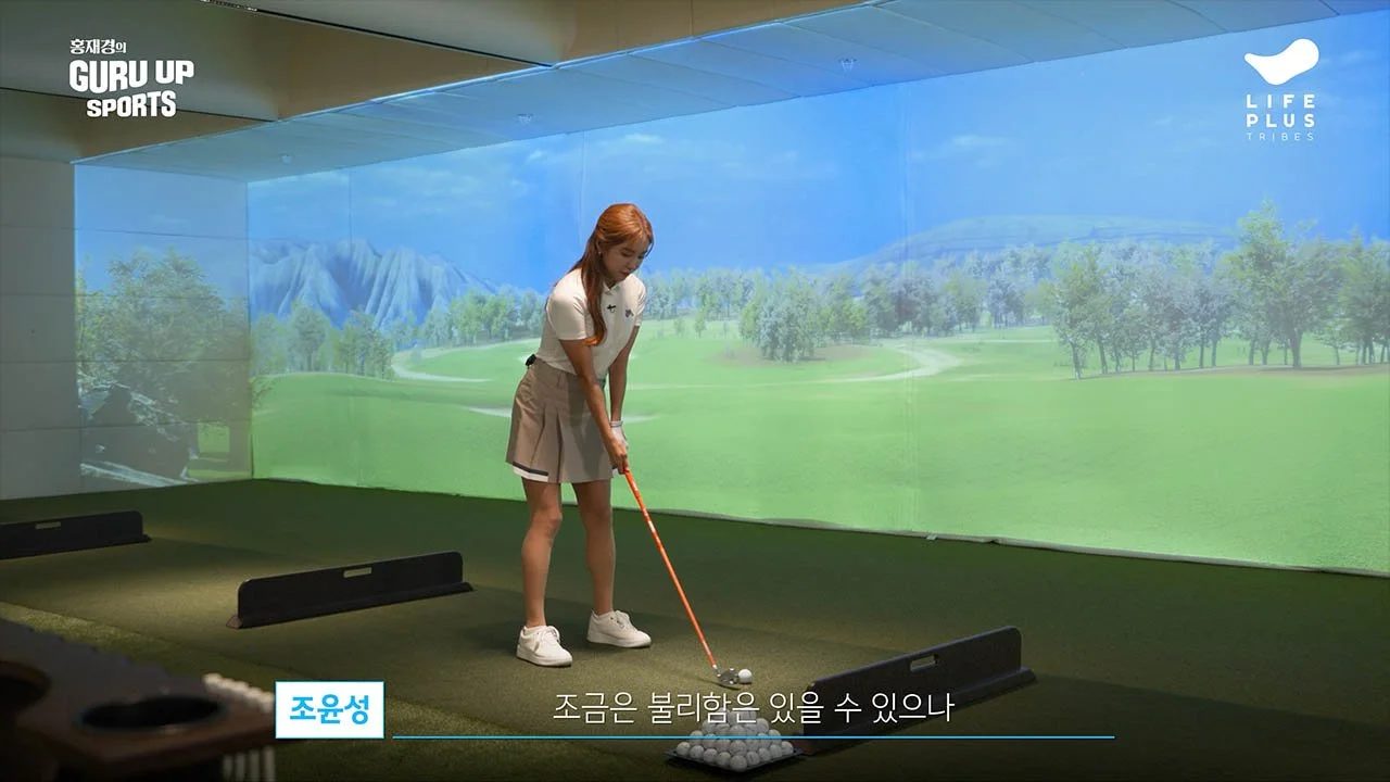 Young woman practicing golf in front of a large screen showing a golf course, in an indoor golf simulation facility.