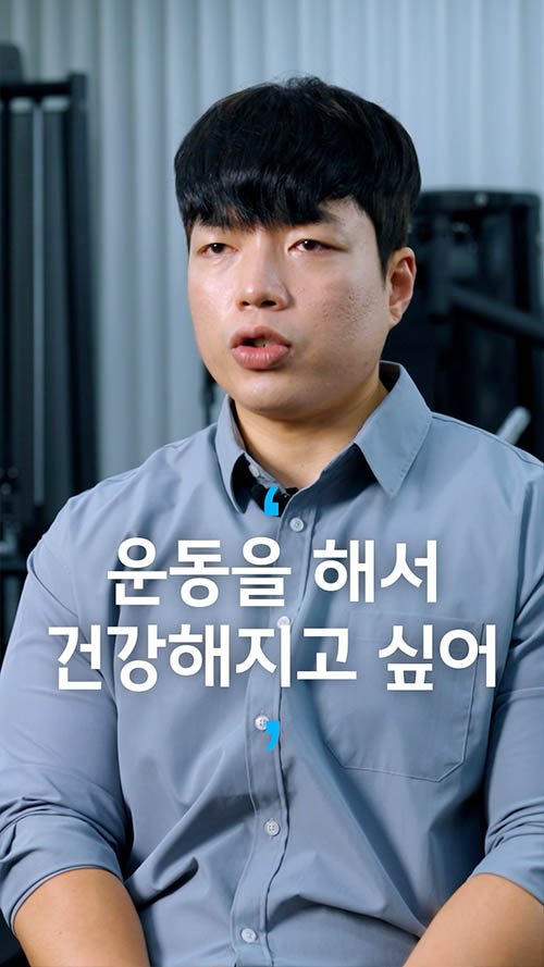 A man with black hair wearing a light gray shirt sitting indoors with a blurred background. Korean text overlaying the image says, '운동을 해서 건강해지고 싶어,' which means, 'I want to become healthy through exercise.'