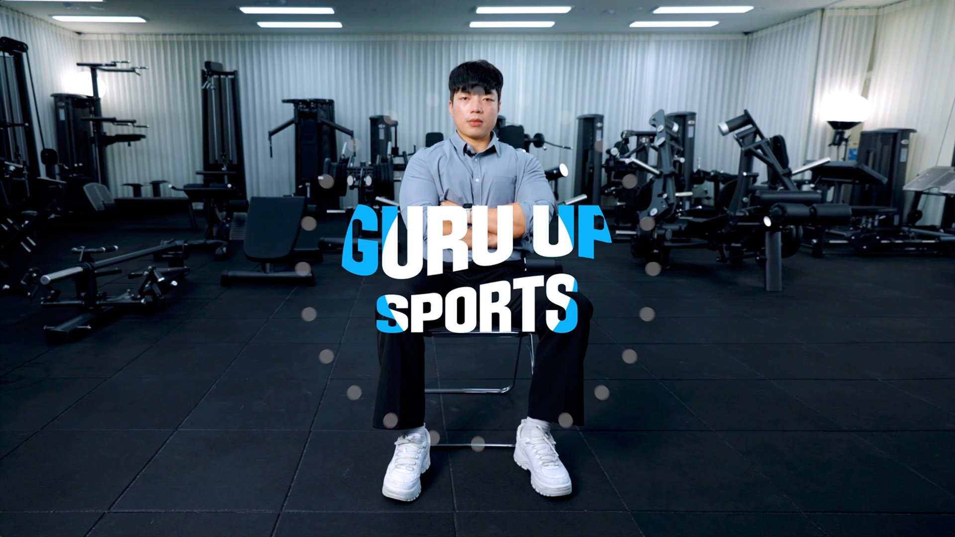 A young man with dark hair and wearing a gray shirt, sitting on a chair with his arms crossed in a gym with exercise equipment in the background and the text 'GURU UP SPORTS' overlaid.