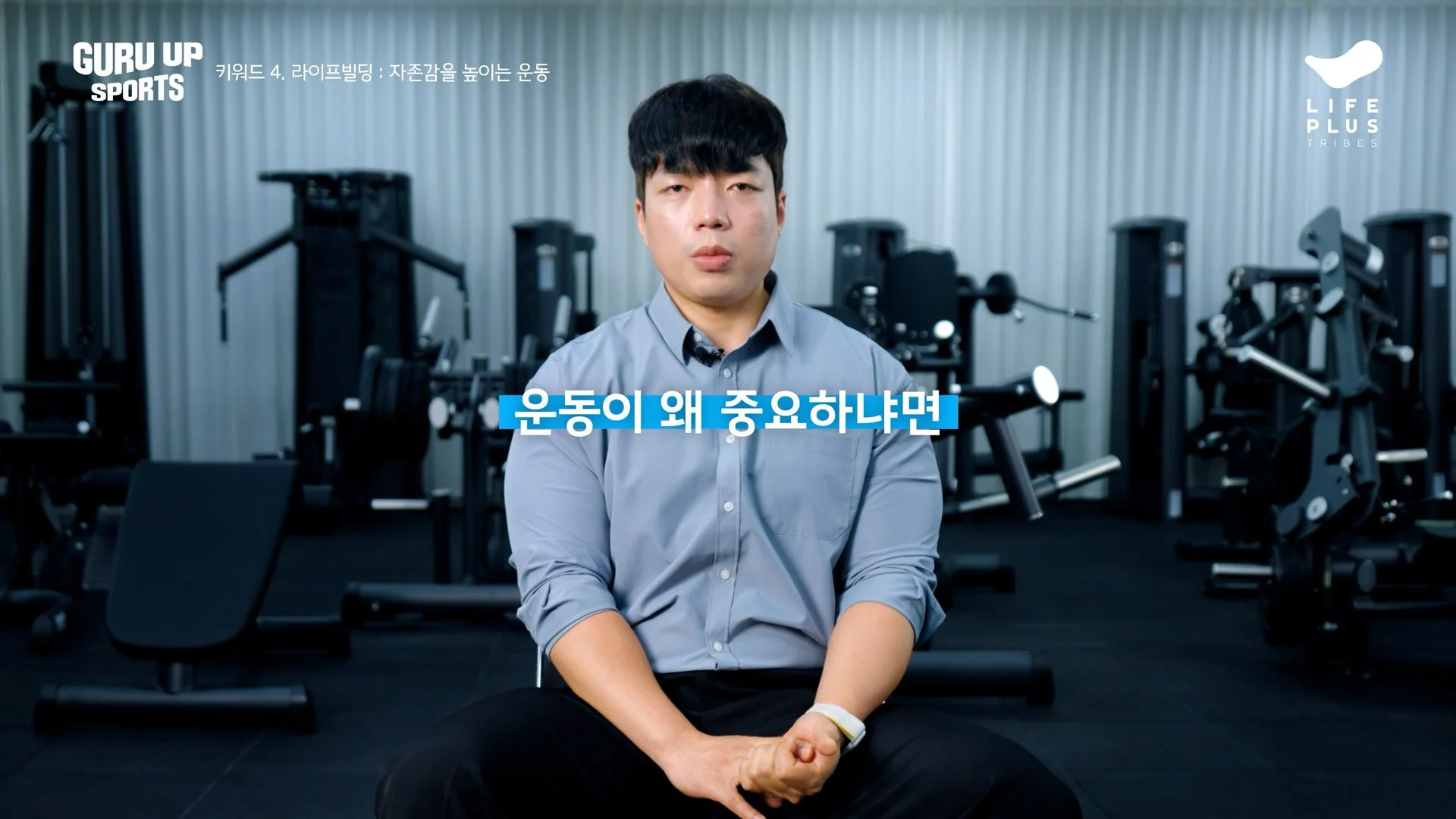 A man with black hair wearing a gray shirt sitting in a gym with workout equipment in the background, with Korean text and a blue banner in the foreground.