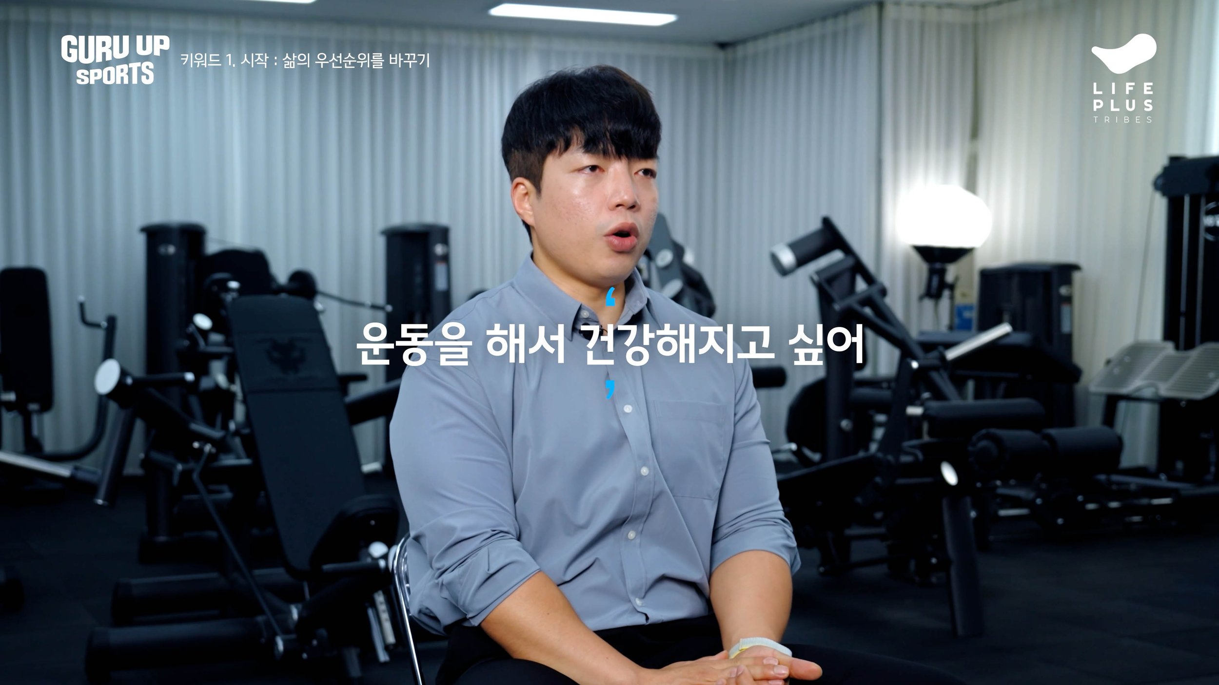 A man sitting in a gym with exercise equipment in the background, speaking with Korean text and a Korean subtitle.