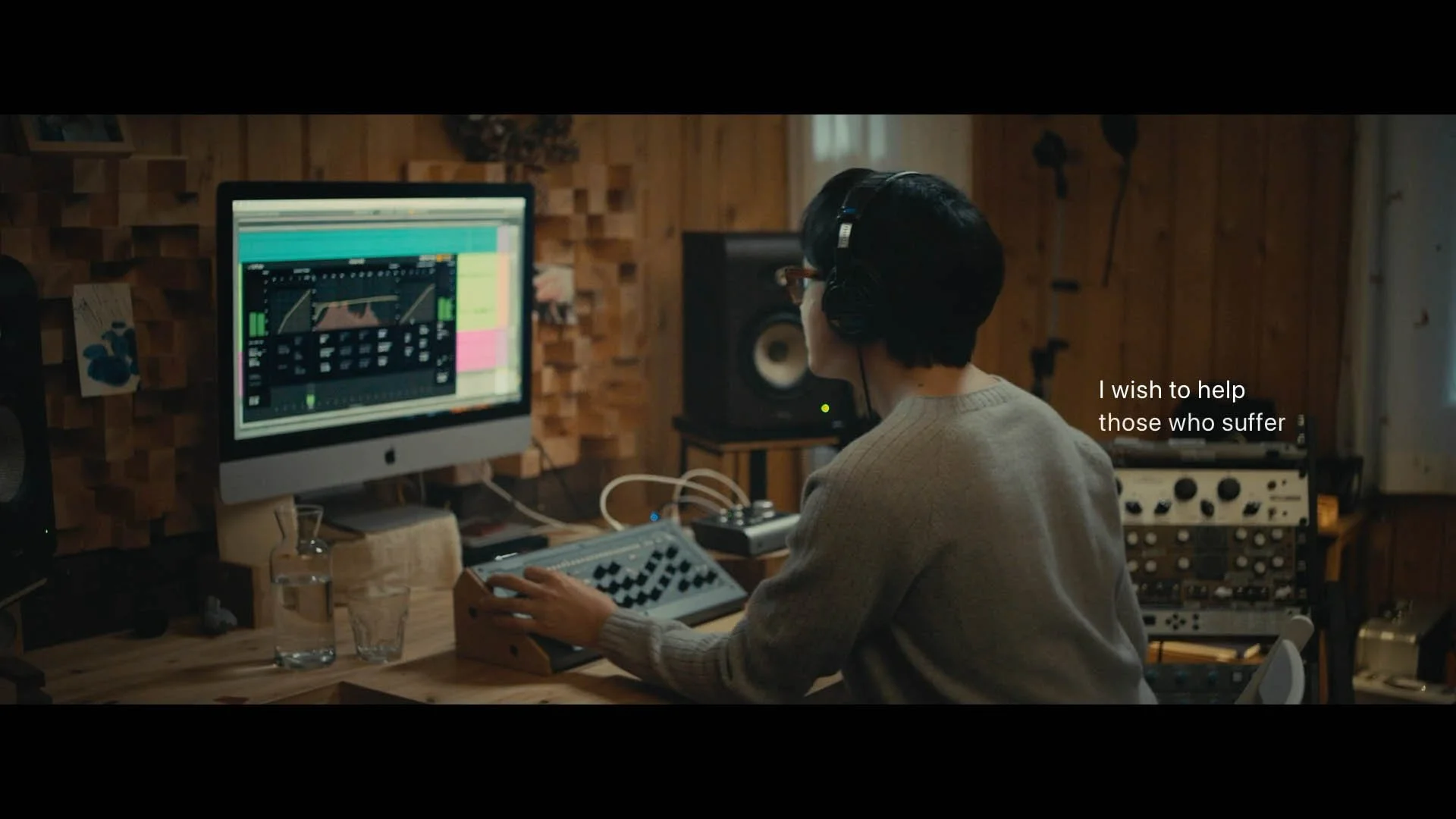 A person in a music studio wearing headphones, working with electronic music equipment, with a computer displaying sound waves, and a subtitle saying 'I wish to help those who suffer'.